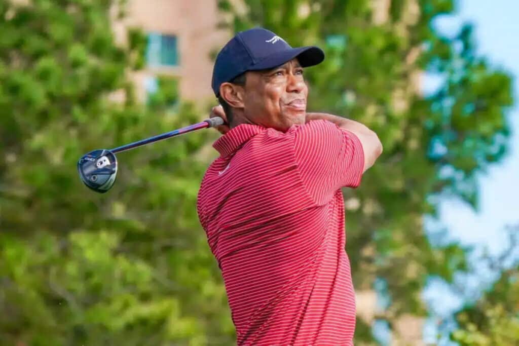 Une photo du golfeur Tiger Woods prenant une photo sur un parcours de golf tout en portant une chemise rouge clair et une casquette de baseball bleu foncé. Les arbres sont visibles flous en arrière-plan