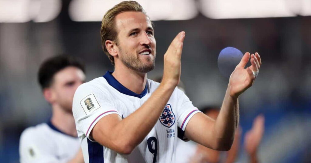Le footballeur anglais Harry Kane joue au football pour l'Angleterre tout en regardant à sa gauche sur un terrain, souriant et applaudissant dans une chemise blanche avec bordure bleue. Un coéquipier est flouement visible en arrière-plan dans le stade