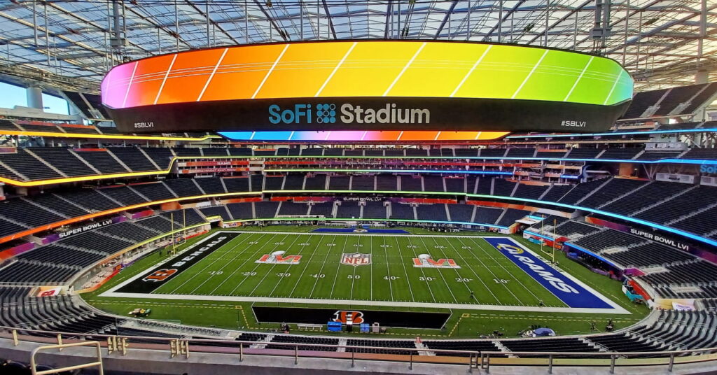 Une photo de la Coupe du monde 2026 et du stade de football américain, le SoFi Stadium de Los Angeles, montrant l'intérieur du stade depuis les sièges en hauteur jusqu'aux côtés avec un grand écran vidéo circulaire montrant des couleurs vives, des rangées de sièges noirs sur du béton et un terrain bordé de vert sous un ciel bleu.