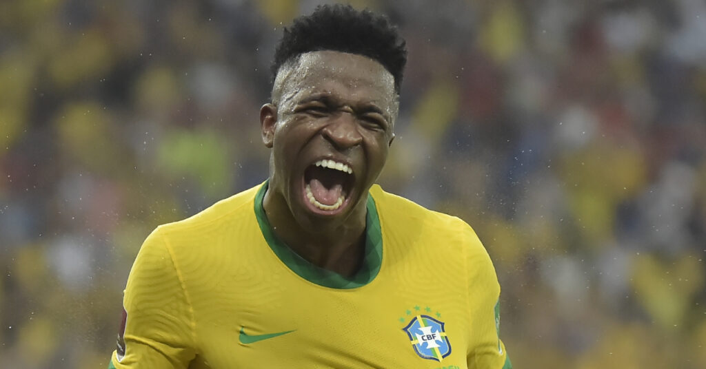 Le footballeur Vinicius Junior crie de fête et sourit tout en portant un maillot de l'équipe nationale de football du Brésil dans un stade. Le maillot est jaune avec une bordure verte et porte une coche Nike verte et un badge Brésil sur la poitrine.