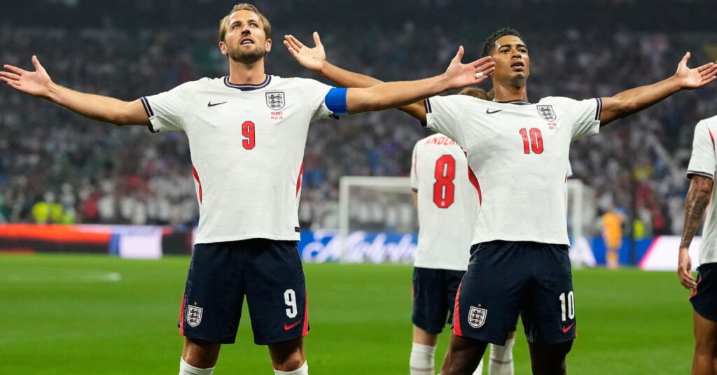 Les footballeurs Harry Kane et Jude Bellingham écartent les bras et regardent vers le haut pendant la Coupe du monde 2026 tout en portant le kit de football de l'équipe nationale d'Angleterre composé d'un maillot blanc avec une bordure bleue et leurs numéros en rouge sur le maillot avec un short bleu foncé et une bordure blanche avec l'insigne de l'Angleterre et leurs numéros en blanc.
