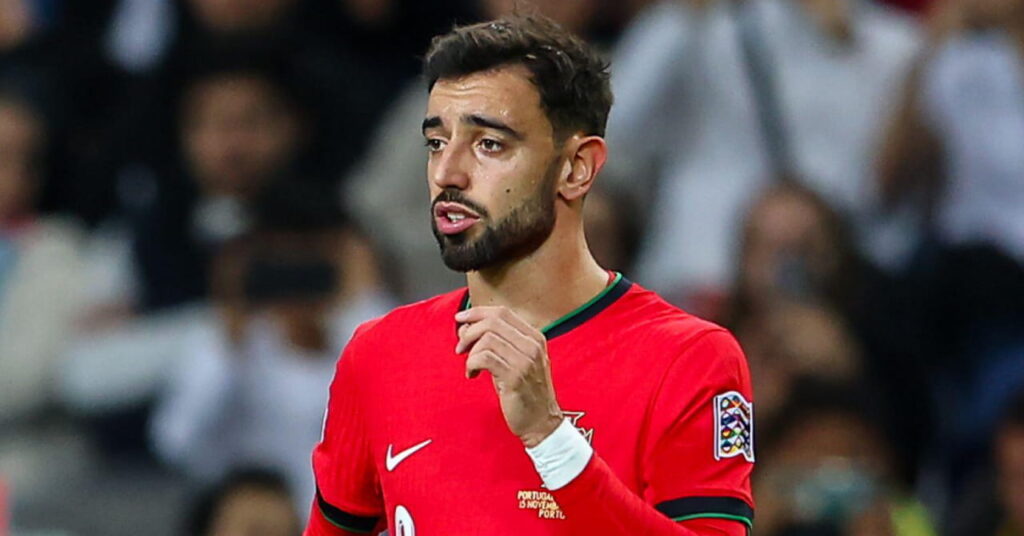Une photo du footballeur Bruno Fernandes regardant à sa droite alors qu'il se trouve sur un terrain de football. Le joueur de football porte un maillot de l'équipe nationale de football du Portugal en rouge avec des bordures vertes et noires et un logo et un numéro Nike blancs sur sa poitrine. Une foule est flouement visible en arrière-plan dans le stade