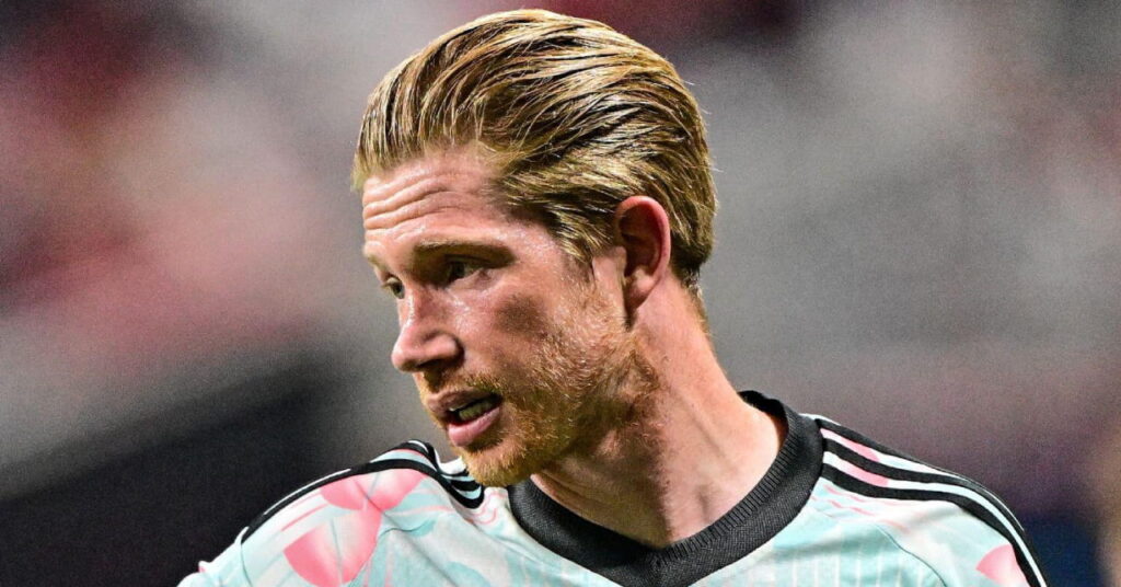 Une photo du footballeur Kevin De Bruyne regardant à sa droite alors qu'il se tenait sur un terrain, souriant et jouant pour l'équipe nationale belge de football, vêtu d'un maillot bleu clair et rose avec un col noir et des lignes noires sur les épaules.