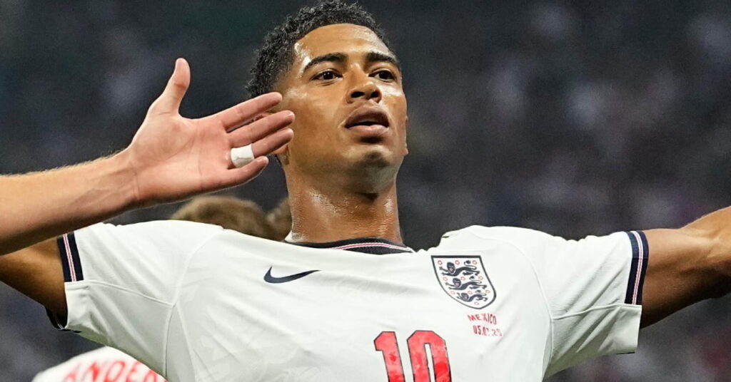 Le footballeur Jude Bellingham écarte les bras et regarde vers le haut pendant la Coupe du monde 2026 tout en portant le kit de football de l'équipe nationale d'Angleterre composé d'un maillot blanc avec bordure bleue et son numéro en rouge sur le maillot avec un short bleu foncé et une bordure blanche comportant l'insigne de l'Angleterre et son numéro en blanc.