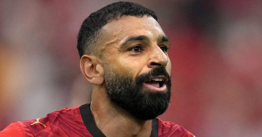 Le footballeur égyptien Mohamed Salah regarde vers sa gauche, la bouche ouverte, alors qu'il se trouve dans un stade de football lors de la Coupe du monde de football de la FIFA 2026 et porte un maillot de football rouge de l'équipe nationale avec une bordure noire.