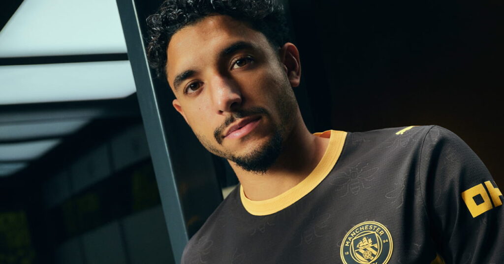 Le footballeur de Man City, Omar Marmoush, modélise le maillot extérieur à prédominance noire du Manchester City Football Club avec une bordure jaune et Etihad Airways sur le devant dans une pièce sombre.