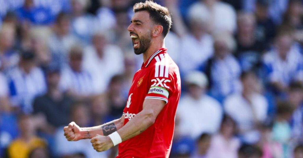 Le footballeur portugais Bruno Fernandes lève les deux bras et regarde vers sa gauche tout en célébrant un but pour l'équipe de Premier League du Manchester United Football Club dans une chemise rouge avec une bordure blanche et noire.