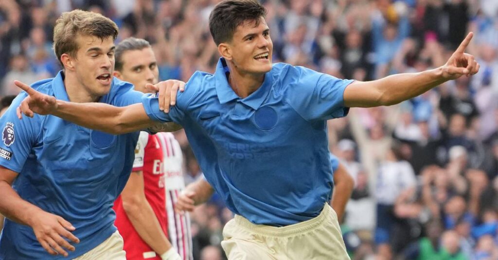 Le footballeur Chema Andres levant les deux bras et ses index pour célébrer un but alors qu'il jouait pour le club de Premier League Brighton et Hove Albion Football Club dans une chemise bleue et un short blanc devant une foule floue visible avec le footballeur croate Luka Vuskovic le félicitant