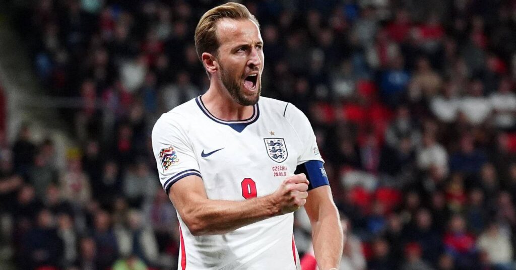 Le footballeur Harry Kane célèbre un but en levant son bras droit et en serrant le poing tout en rugissant et en regardant vers sa gauche tandis que le joueur de l'équipe de Bundesliga du Bayern Munich Football Club joue dans le maillot domicile de l'équipe nationale de football d'Angleterre de couleur blanche avec des détails bleu foncé avec le logo Nike et l'écusson des Trois Lions sur la poitrine au-dessus de son numéro neuf en rouge avec une foule floue visible derrière lui.