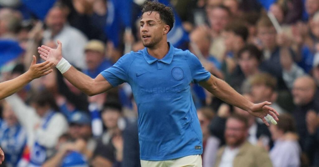 Le footballeur grec Charalampos Kostoulas lève la main droite pour célébrer un but alors qu'il joue pour l'équipe de Premier League Brighton and Hove Albion Football Club dans une chemise bleue et un short blanc avec une foule de stade floue visible en arrière-plan et un plâtre sur le quatrième doigt de sa main gauche tendue