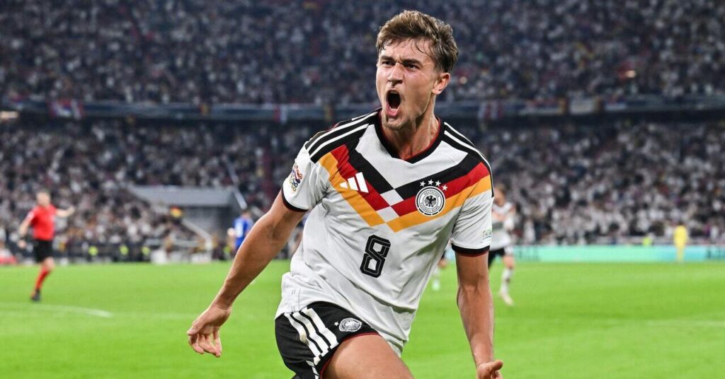Le footballeur Tom Bischof rugit pour célébrer un but tandis que le joueur de l'équipe de Bundesliga du Bayern Munich Football Club joue dans le kit domicile de l'équipe nationale d'Allemagne composé d'une chemise blanche avec des détails noirs, rouges et jaunes avec les logos Adidas et l'association et son numéro huit en noir sur la poitrine et des chemises noires avec bordure blanche sur un terrain d'herbe verte devant une foule