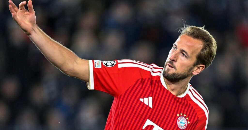 Le footballeur anglais Harry Kane lève le bras droit et déplace la tête vers la gauche tout en se baissant légèrement et en jouant pour l'équipe de Bundesliga du Bayern Munich Football Club dans une chemise rouge avec des détails blancs et l'insigne du club, le logo Adidas blanc et cinq étoiles dorées visibles sur la poitrine. Le logo du ballon de l'UEFA Champions League est visible sur sa manche droite.